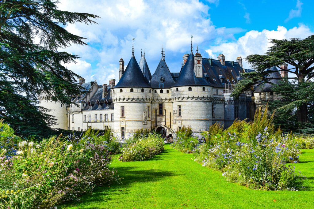 Photo of Château-de-Chaumont showing the castle set against a blue sky with puffy white clouds. In front is a verdant lawn, planted with wildflowers in shades of pink, blue, and lavender. Evergreens flank each side of the photo frame. The scene is bucolic and stately.